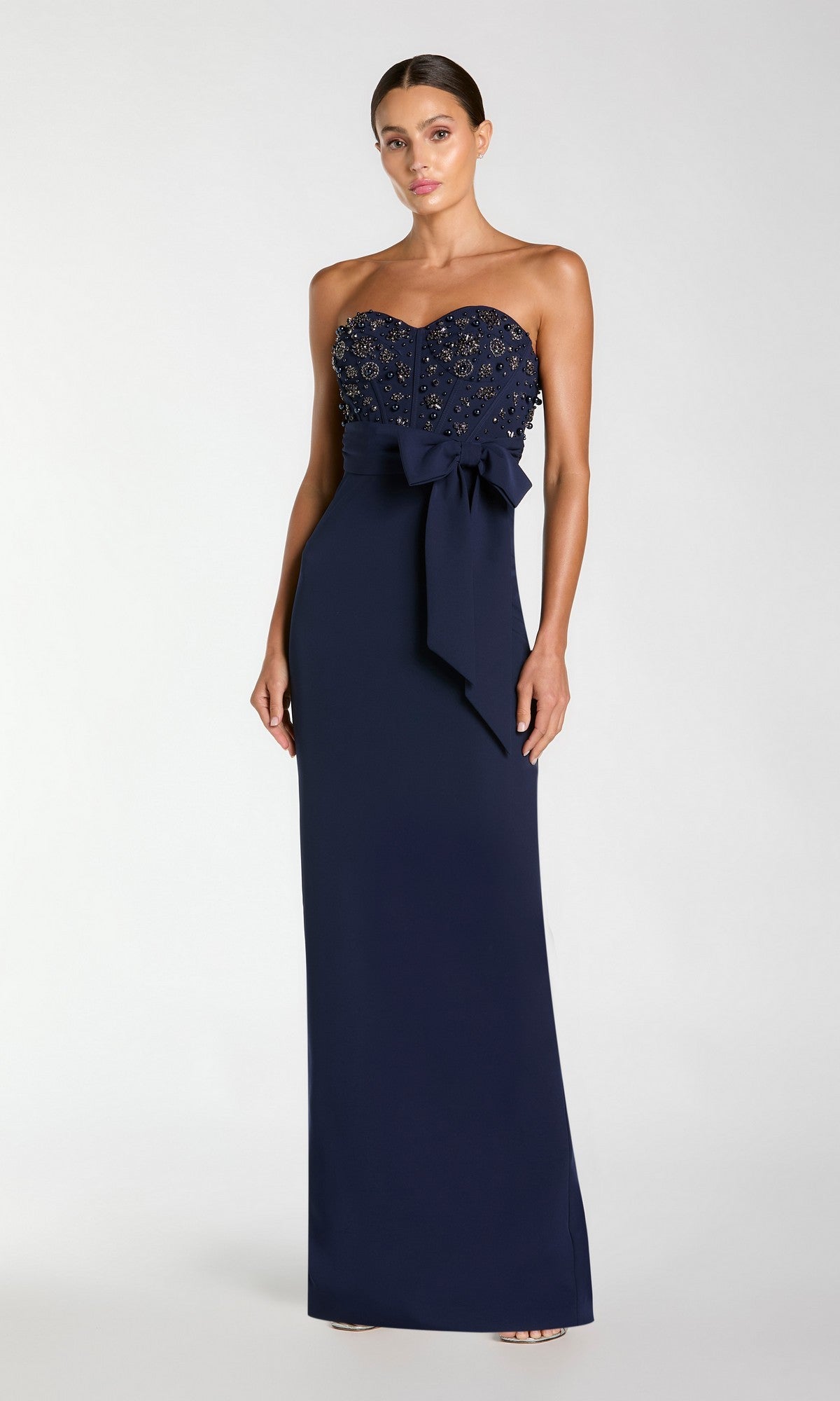 A woman stands against a plain light background wearing the Mac Duggal 2395 Long Formal Dress in Midnight, featuring a strapless design with bead embellishments on the bodice and a bow at the waist.